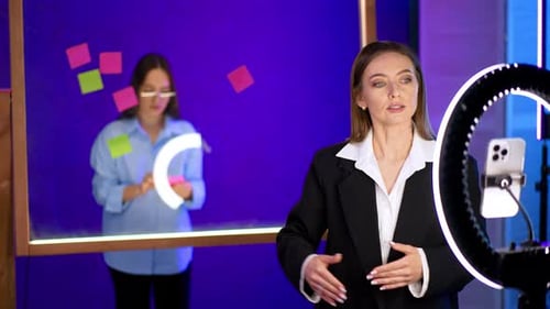 Good-looking Caucasian lady in black jacket takes a video and points at the glass wall behind her.