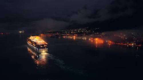 Aerial drone view of a cruise ship moving slowly through a dark and misty bay at night, with faint l