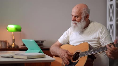 Curieux vieil homme caucasien assis avec une guitare jouant en train de regarder une leçon en ligne sur les réseaux sociaux sur