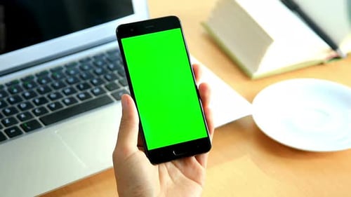 Hand Holds Smartphone with Green Screen at Desk
