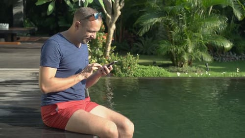 Young man enjoys tropical holiday texting on smartphone by luxury swimming pool