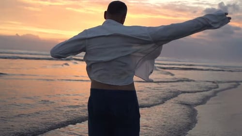 Man Putting on Shirt and Stretching Arms on Beach During Sunset Admire
