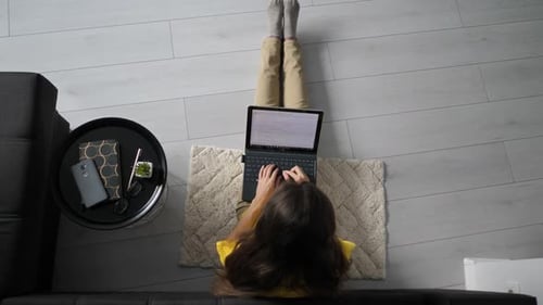 Top view of female freelancer sitting on floor near sofa at home and working on laptop computer.