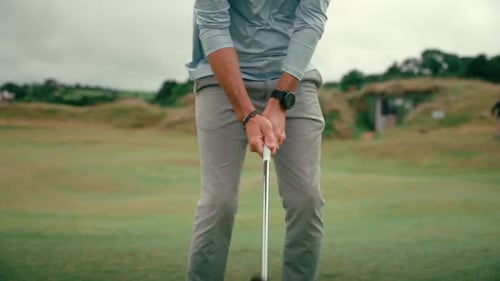 Male golfer sets up shot, lines up wedge club behind ball for chip shot on rolling hilly fairway of