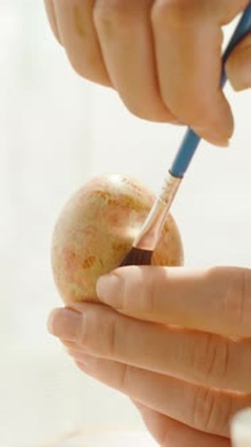 Vertical video. Woman delicately painting a pastel Easter egg, adding fine details with a brush.