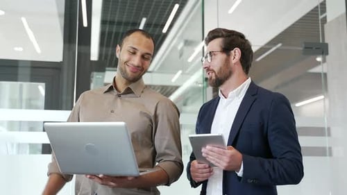 Two smiling businessmen communicate standing in business office. Male colleagues talking during