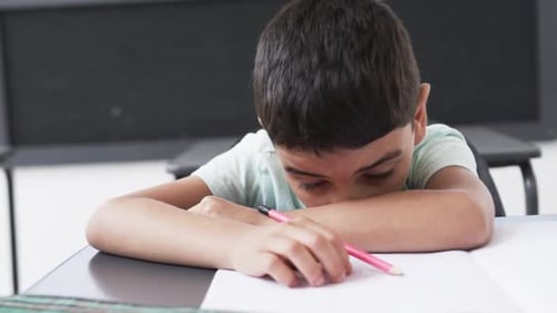 In a school classroom, a young boy rests his head on his arms