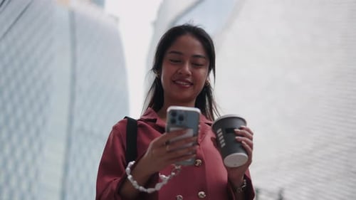 Young Asian Manager Using Smartphone and Drinking Coffee Near an Office Building