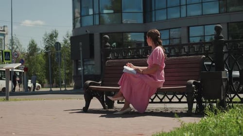 Woman in pink dress sits on park bench writing
