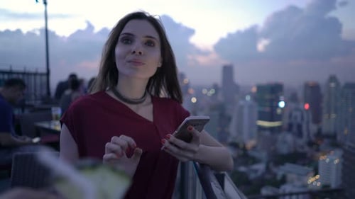 Young Woman with Smartphone Drinking Cocktail Standing on Terrace in Bar Alcohol