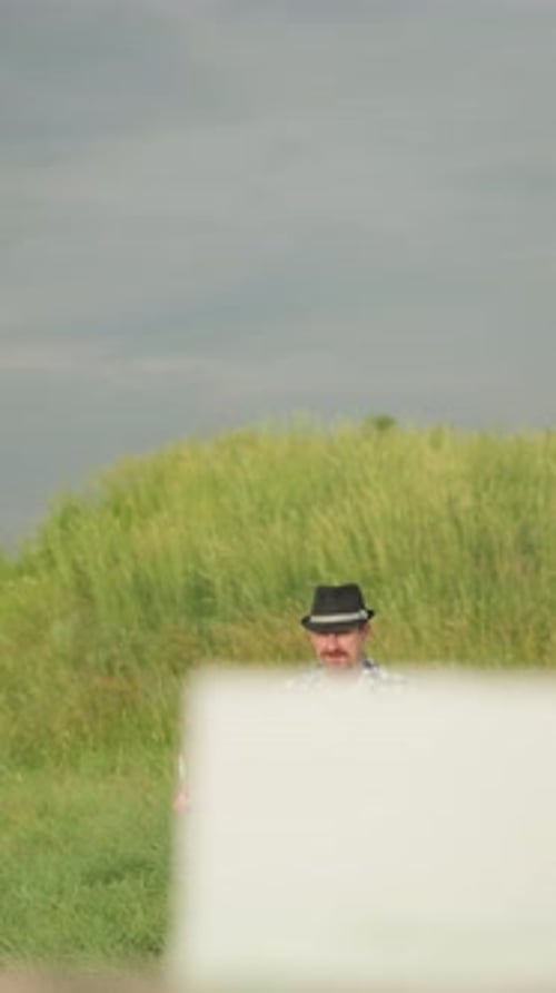 Man Holding Brush Seated in Sunlit Grassy Field Wearing Black Hat