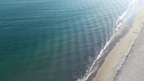 Drone footage of ocean waves crashing gently against the beach at sunset with people walking on the