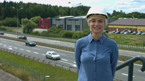 Woman on construction site wearing a hard hat standing above a freeway/highway with a big smile