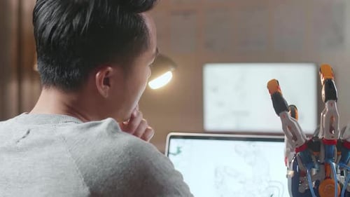 Over The Shoulder View Of A Male Thinking While Designing A Cyborg Hand On A Laptop At Home