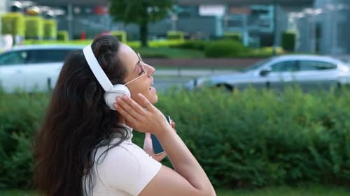 Side View of Beautiful Joyful Woman Listening to Music in White Headphones with Smartphone in Hand