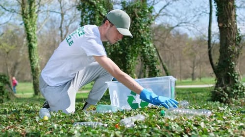 Man collecting plastic garbage in container in a polluted park. Rubber gloves, recycling signs on T-
