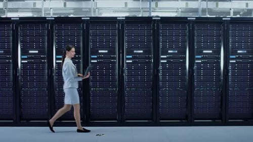 Female Data Center IT Engineer Walking Through Server Rack Corridor with a Laptop Computer. She is