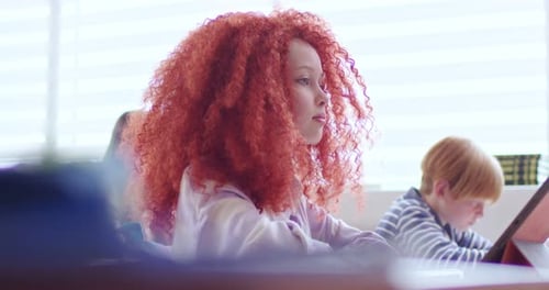Side View of Redhead Schoolgirl Sitting at Desk with Tablet in Bright Classroom Unrecognisable