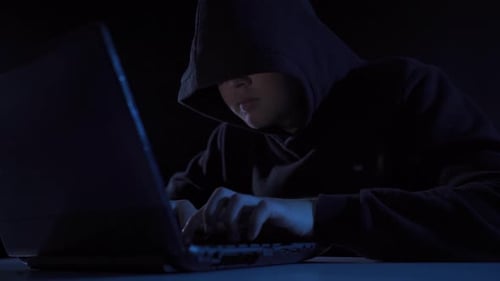 Hooded Figure Typing on Laptop in Dark Room