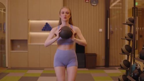 Fitte junge Frau mit langen Haaren in Sportkleidung, die Kniebeugen mit einem schweren Medizinball während eines