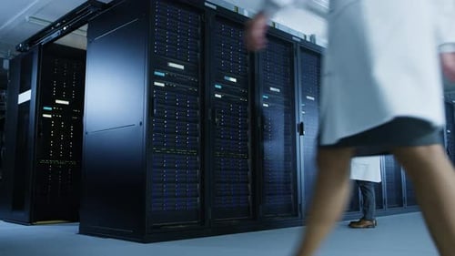 In Data Center: Male IT Technician Wearing White Coat Stands at Open Server Rack Cabinet, Uses Lapt