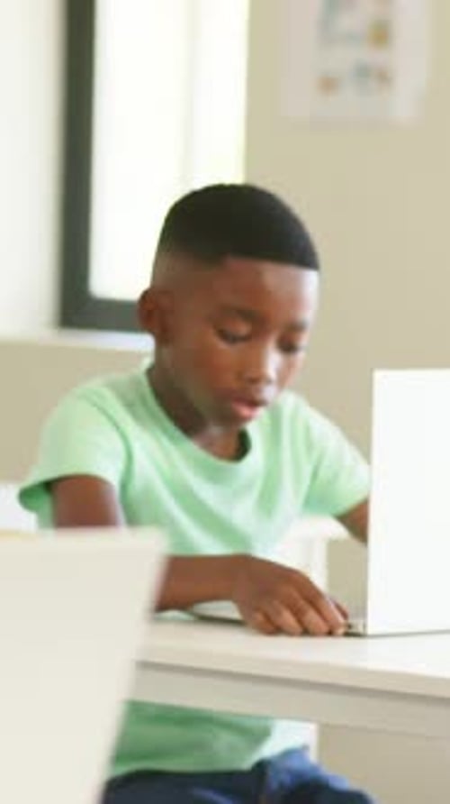 Vertical video: Typing child in green tee on laptop in class, finishing task and signaling teacher