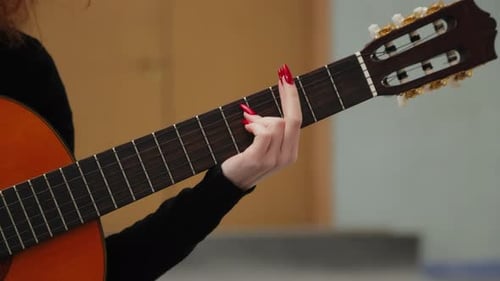 Close-up video of a woman playing chords on an acoustic guitar while sitting on a stool in a room.