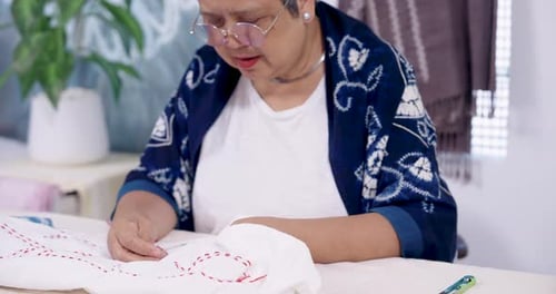 Un artesano asiático de alto nivel se inclina hacia adelante, comprueba las líneas marcadas y luego perfora la tela con una aguja para continuar