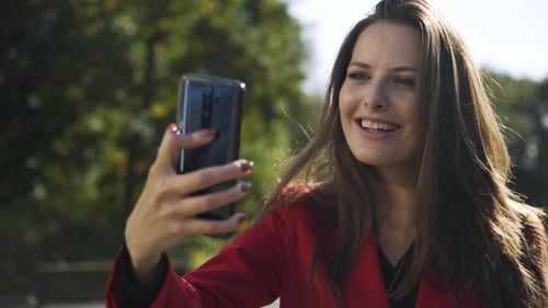 A Young Beautiful Caucasian Businesswoman Takes Selfies with a Smartphone in a Park on a Sunny Day