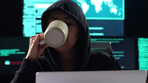 Asian Female Hacker In A Hood Drinking Coffee While Works On A Laptop With Multiple Monitors