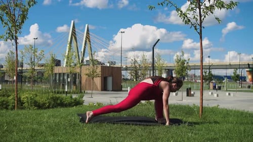 An Adult Woman in Red Training Suit Doing Stretching Exercises in the Park By the Bridge