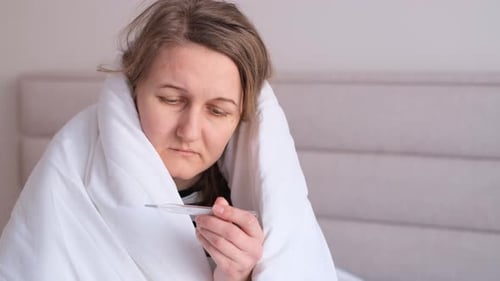 Sick Woman in Bed Looks at Thermometer