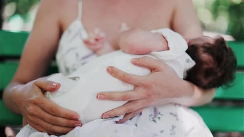 A mother siting on a bench in the park, breastfeeding her baby while enjoying a quiet, peaceful mome