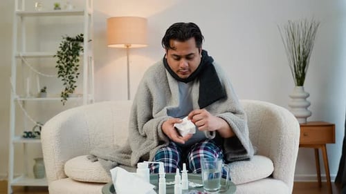 Indian Man with Runny Nose Sits on the Couch at Home Under a Plaid Sneezing in a Napkin