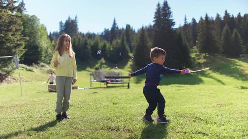 Older Sister Plays with Younger Brother and Teaches Him How to Blow Soap Bubbles on the Backyard in