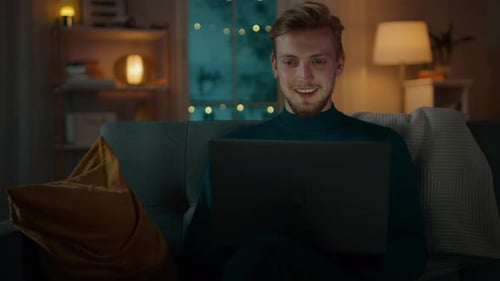 In the Evening Handsome Young Man at Home Sitting on a Couch in the Living Room Works on a Laptop C