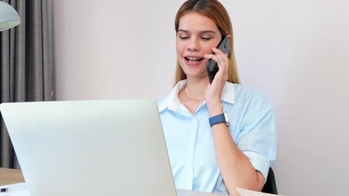 Young caucasian businesswoman talking with smartphone and working on laptop computer in home office.