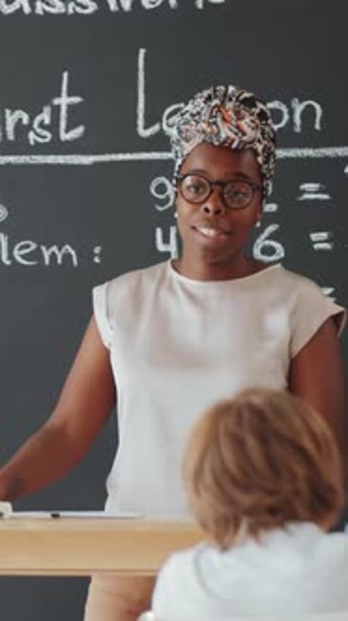 Black Female Teacher Talking at Blackboard during School Lesson