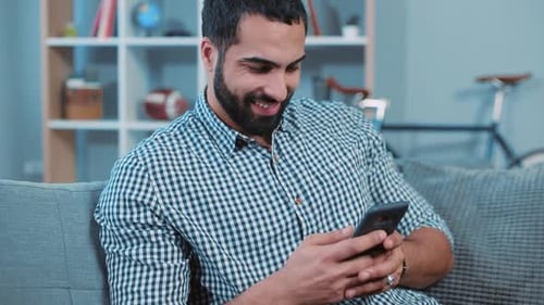 Attractive Young Man Hold Use Phone Smiling Feel Happy Sit on Sofa Home Portrait Close Up Smartphone