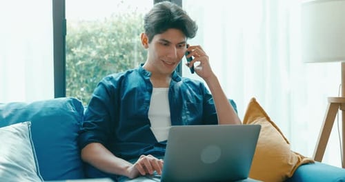 Asia male use laptop and talk on cell phone remote working on sofa in living room at home office.