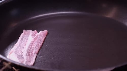 Raw bacon strips are placed into fry pan by Caucasian right hand