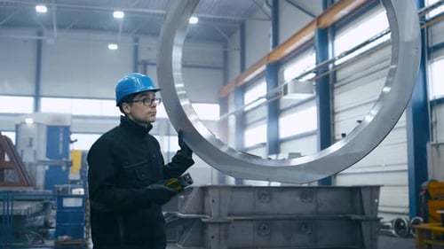 Factory worker in a hard hat is moving a metal detail with a remote crane.