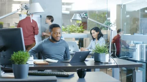 Busy International Office, African-American Man Working at His Desk on a Laptop, He Writes in Remin