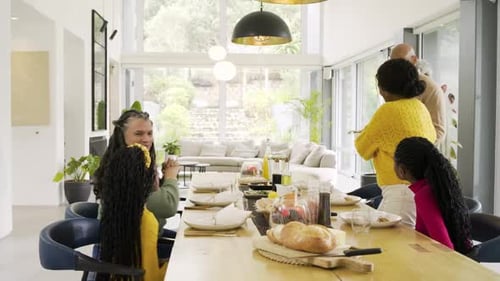 Diverse family greeting arriving grandpa, hugging, passing dishes at dining table to share meal