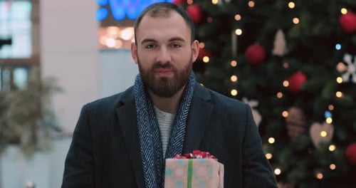 Portrait of Intelligent Man Gives Christmas Present Stand Look at Camera Smiling Near Christmas Tree