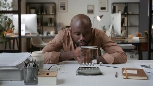 Portrait of Black Man Resting Head on Fists at Office Desk with Newtons Cradle
