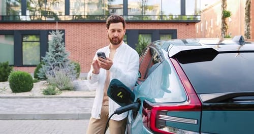 Man uses phone while charging electric vehicle outdoors