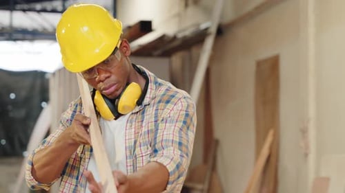 African American Carpenter Inspecting a Piece of Wood While Carrying a Wooden Plank in a Workshop