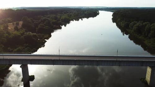 White truck with a red semi-trailer drives on a highway bridge over a river during sunset.