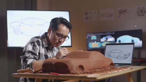 Asian Man Designer Looking At A Laptop And Using Rake Or Wire To Smooth Out The Surface Of Car Clay